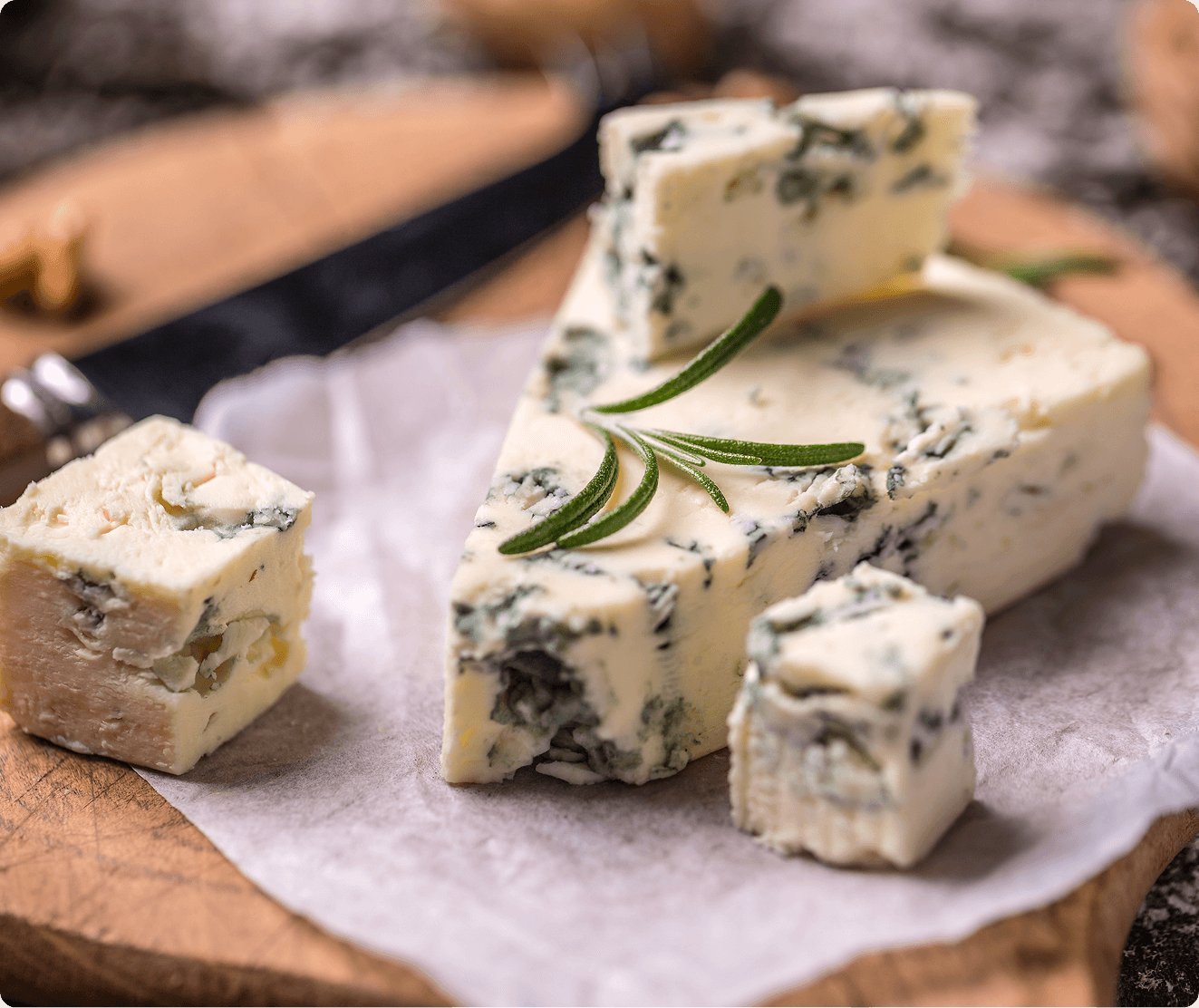 GettyImages-1306438018 A wedge of blue cheese with rosemary garnish sits on parchment paper, surrounded by small cheese cubes on a wooden board. A knife is in the background.