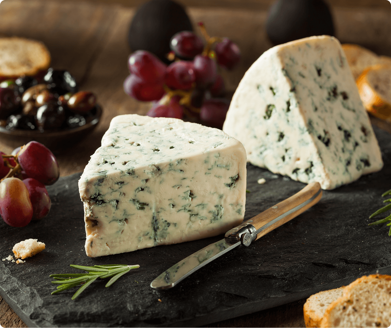 Two wedges of blue cheese on a slate board with a cheese knife, surrounded by red grapes, olives, rosemary, and bread slices.