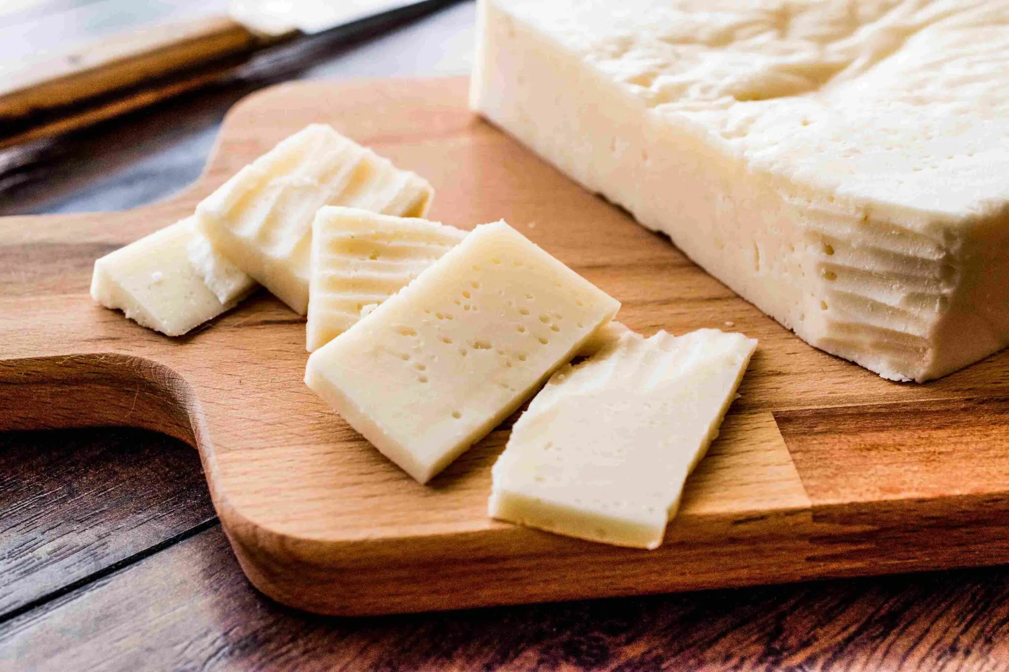 GettyImages-806399918_11zon A block of cheese with several slices cut on a wooden cutting board, placed on a dark wooden surface.