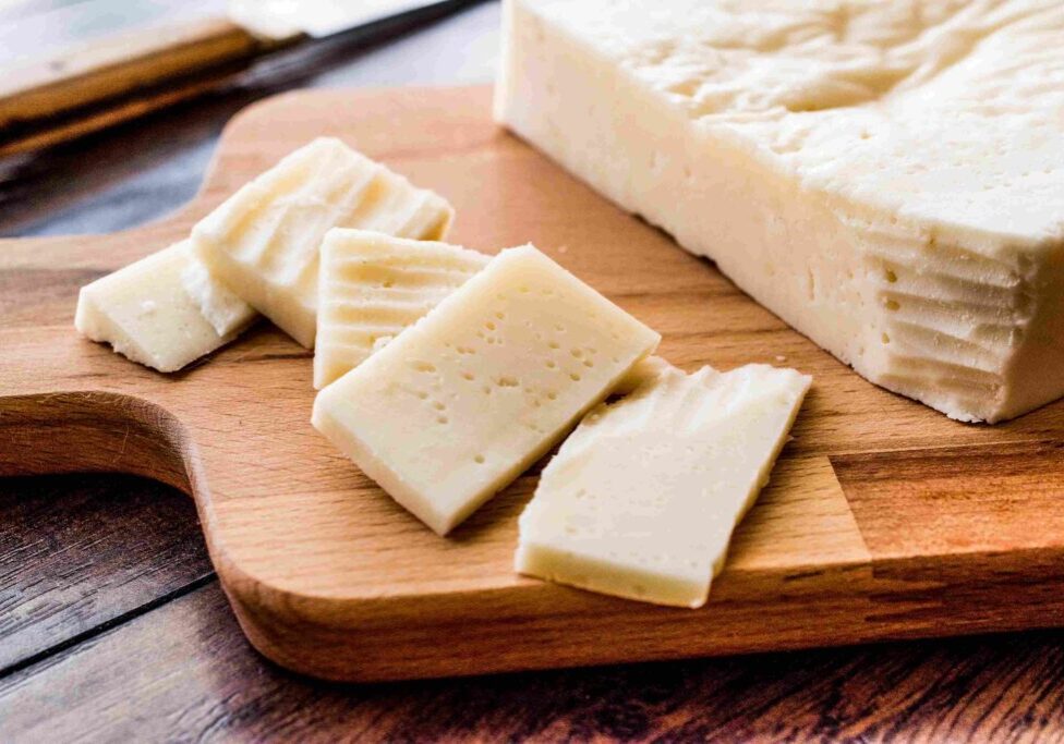 A block of cheese with several slices cut on a wooden cutting board, placed on a dark wooden surface.